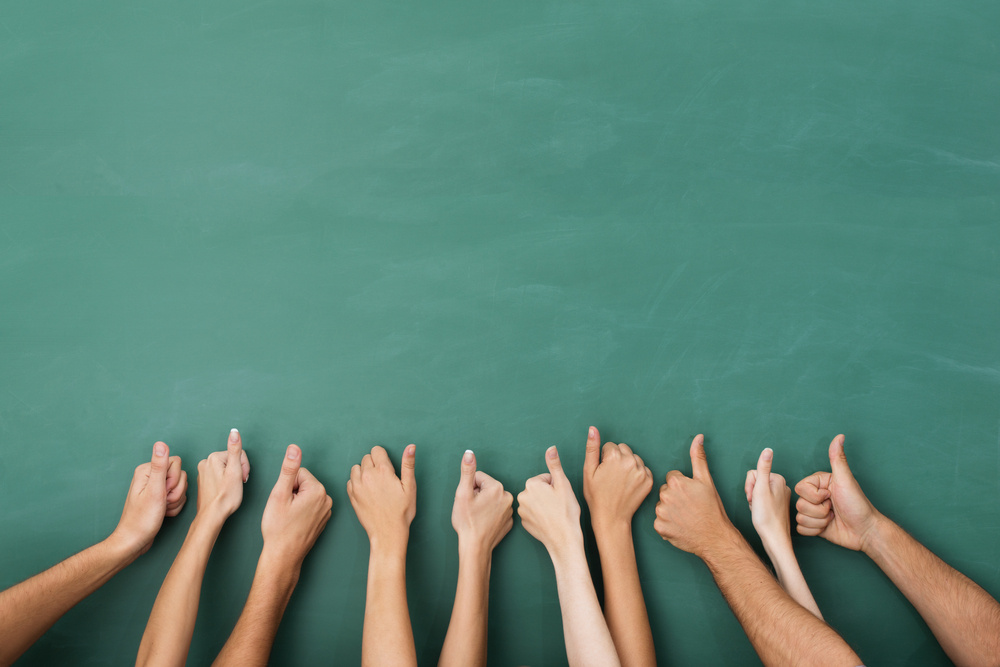 Close up view of the hands of a group of people giving a thumbs up gesture of approval an success with their hands raised against a blank green chalkboard with copyspace-2