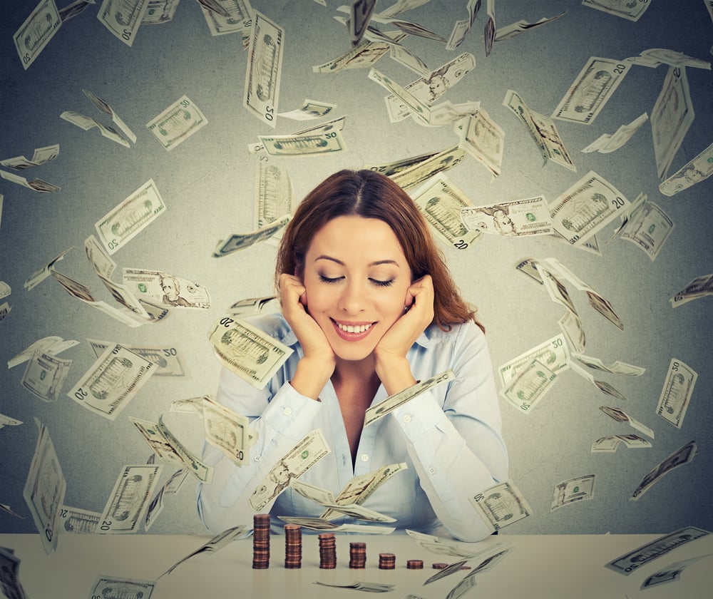 Excited happy young woman sitting at table with growing stack of coins under a money rain isolated on gray wall background. Positive emotions financial success luck good economy concept