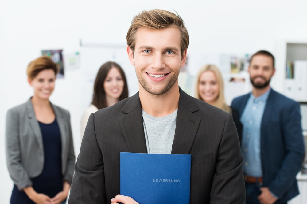 Successful smiling young male job applicant holding a blue file with his curriculum vitae posing in front of his new work colleagues or business team-2